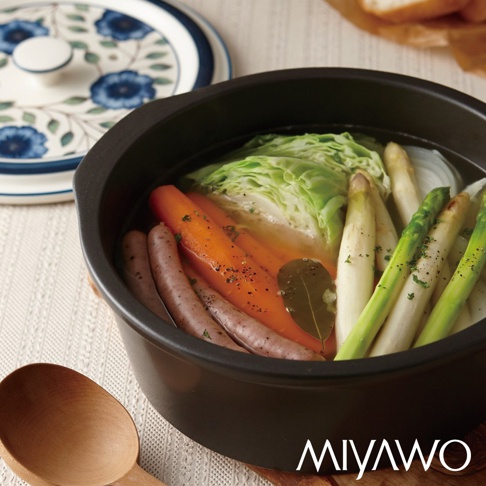 黑色陶土湯鍋內盛有香腸、胡蘿蔔、高麗菜、蘆筍和洋蔥，湯頭清澈，鍋蓋上有藍色花卉圖案。