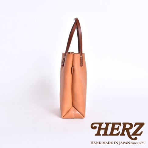 從側面角度拍攝的日本HERZ職人手作皮革托特包，呈現淺棕色皮革材質與深棕色提把的對比，包身線條簡約俐落，展示了底部與側面的精緻縫線細節，右下角印有HERZ品牌標誌。
