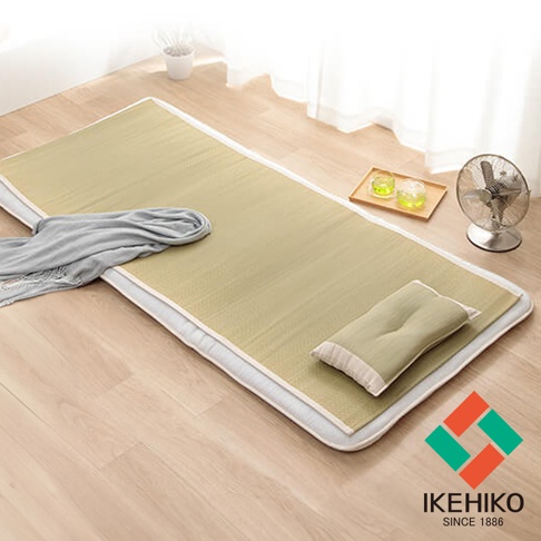 淺棕色藺草編織的單人涼蓆，鋪設在木質地板上，蓆面上有灰色毛巾和一個淺綠色枕頭，角落有品牌標誌。