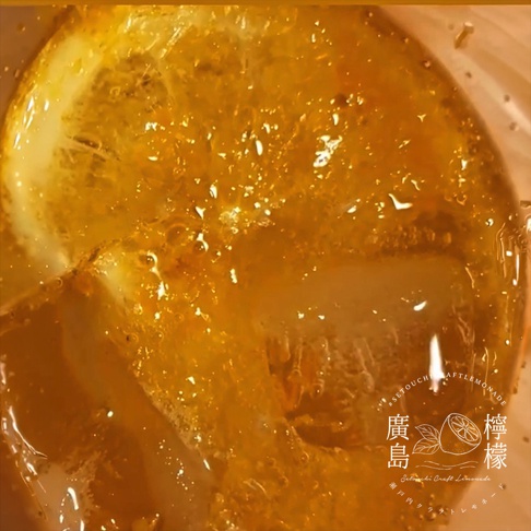 這張由上方俯視的特寫照片中，一杯清涼的廣島手工蜜柑檸檬釀充滿了氣泡，琥珀色的液體映照出光影，杯中漂浮著一片新鮮檸檬切片與幾塊晶瑩剔透的冰塊，右下角印有標示著廣島檸檬與 Setouchi Craft Lemonade 字樣的白色品牌圓形標章。