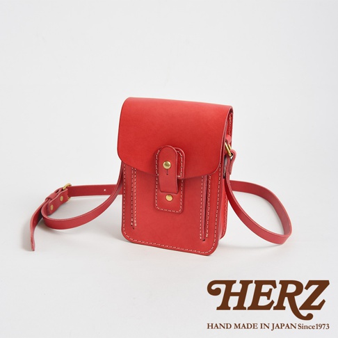 紅色皮革製成的職人手作休閒散步包，帶有可調節長度的皮革背帶，包蓋處有雙重按扣設計，包身側面有縫線裝飾。