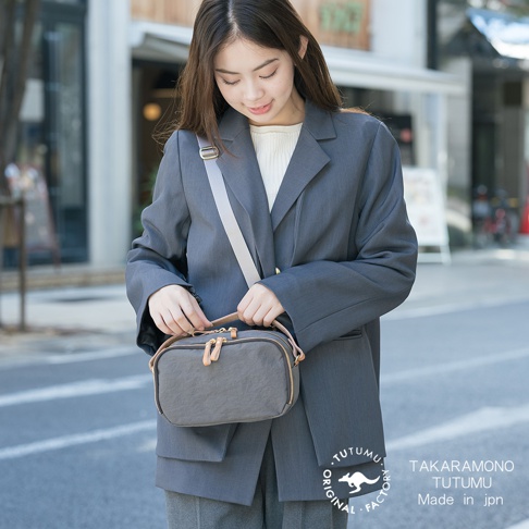 一位年輕女子穿著灰色西裝外套，手提一個灰色兩用斜背包，包上有兩個拉鍊和皮革裝飾。
