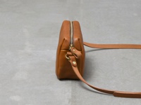 職人手作皮革Organ輕巧拉鍊單肩包，採用棕色皮革製成，配有金色拉鍊和金屬配件，展現簡約俐落的設計。