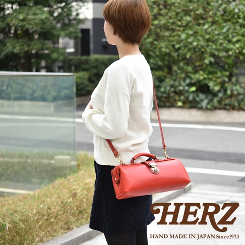 日本HERZ手工皮革橫長形杜勒斯單肩包，咖啡色皮革包身搭配紅色肩帶，包款設計簡約，可在右下角看到品牌標誌。