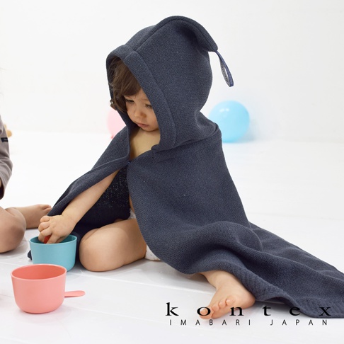 一個穿著深藍色針織風純棉連帽浴巾的嬰兒，坐著玩水杯。浴巾上有一個吊牌，背景有粉色和藍色氣球。