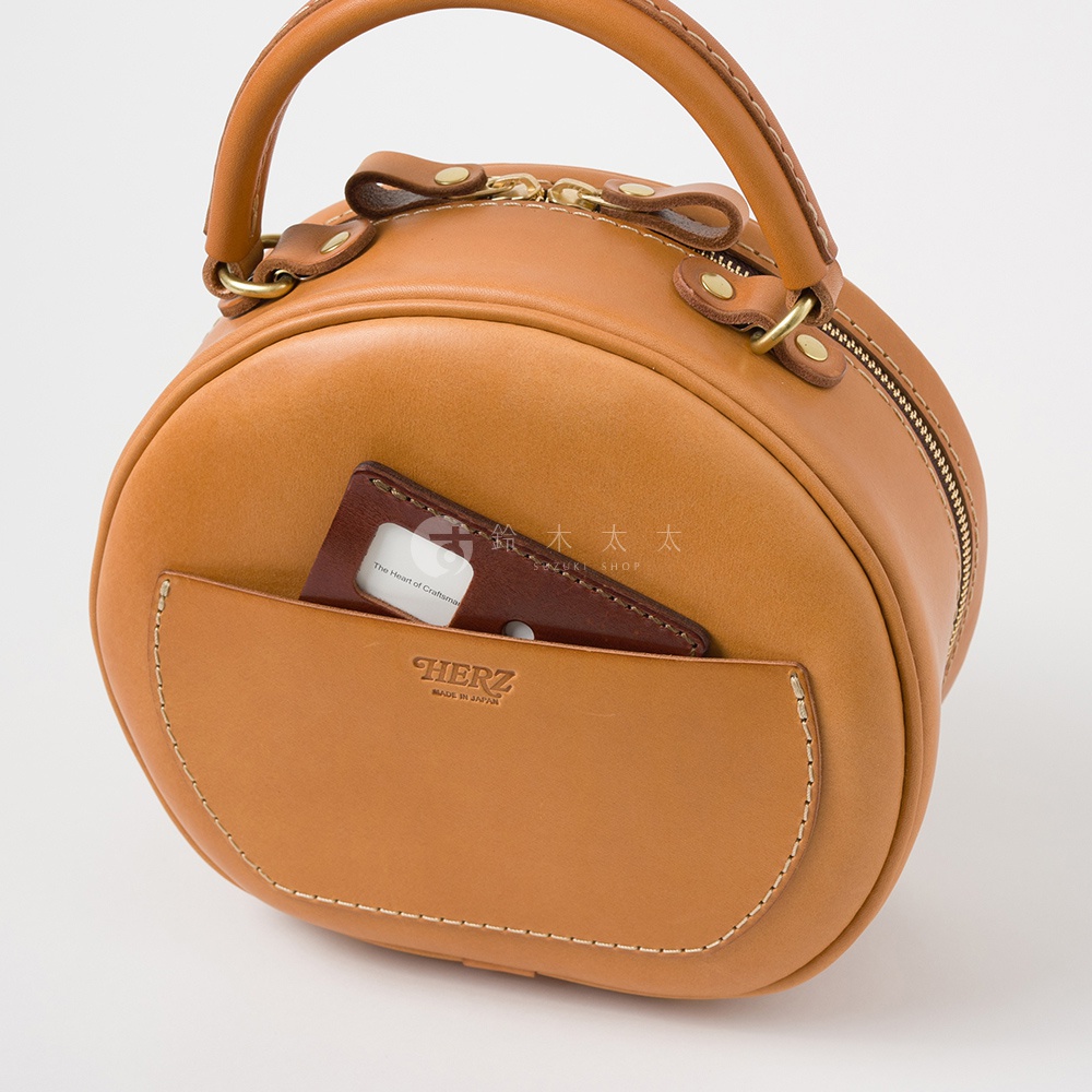 日本HERZ 職人手作皮革兩用圓形包 Q-13，圓潤的棕褐色皮革主體，正面設有可置放卡片的口袋，上方有拉鍊開合，搭配一條可拆卸的圓形提把，整體散發職人手作的質感。