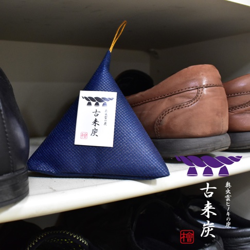 放在鞋櫃層架上的三角形藍色除濕包，表面呈現細緻的紋理質感，頂端附有金色掛繩。包裝正面貼有寫著古來炭的白色方形標籤，並印有繪馬形狀的圖騰，右下角則標示著奧出雲檜木炭的文字與圖案，四周環繞著幾雙男士皮鞋。