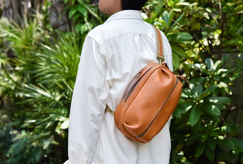 棕色皮革製成的立體運動斜背包，配有兩條拉鍊，肩帶和包身都有縫線裝飾，斜背於穿著白色襯衫的人身上。