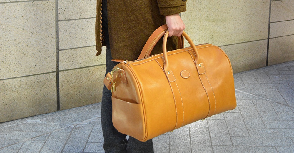 職人手作皮革肩背手提拉鍊波士頓包，呈現溫潤的焦糖棕色，選用光滑皮革材質，搭配白色縫線工藝，包身兩側設有側袋，質感細節豐富。