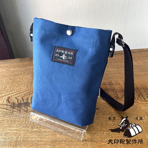 藍色帆布平板收納側背包，包身上有一個印有日文文字和圖案的方形布標，右側有黑色可調整長度的背帶。