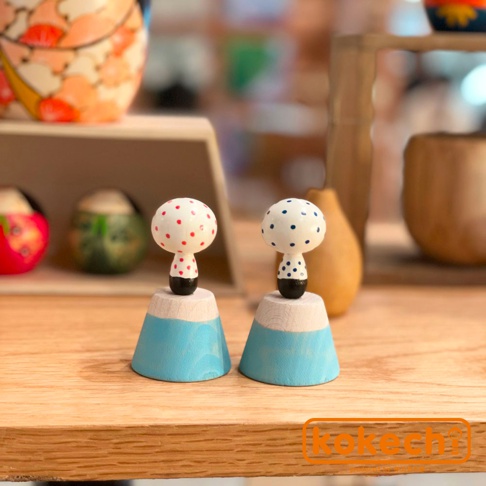 兩個木芥子開運擺飾，一個頭部有紅點，一個頭部有藍點，皆立於淺藍色富士山造型底座上，材質為木頭。