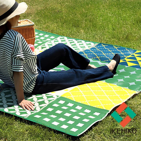 防水 PP 戶外野餐墊，由綠色、黃色、藍色和白色色塊拼接而成，印有幾何圖案，一人坐在墊上，背景為草地。