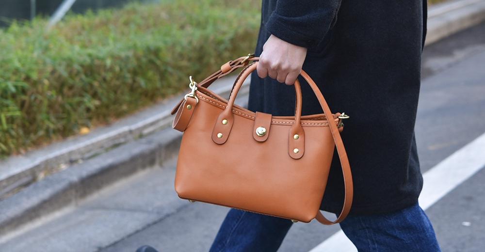 一手提著棕褐色皮革迷你托特包，包上有金色金屬釦和縫線細節，另有可拆卸斜背帶。