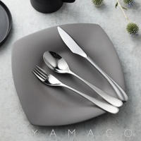 霧面不鏽鋼晚餐叉、湯匙和刀子，擺放在灰色的盤子上，背景有模糊的黑色杯子和裝飾性的植物。