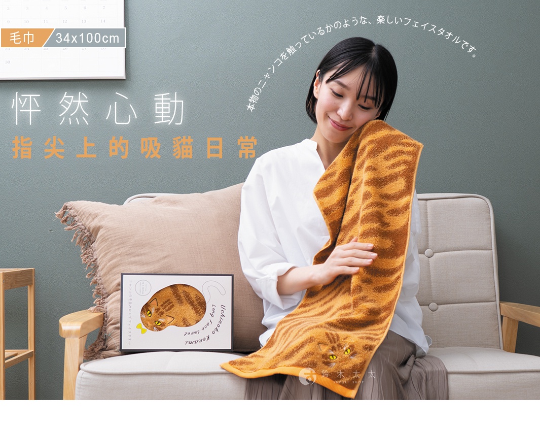 一位女性坐在淺色沙發上，手中輕撫著一條印有橘色虎斑花紋的長方形毛巾，毛巾邊緣繡有可愛的貓咪臉部圖案。她身旁放著印有相同貓咪圖樣的商品包裝盒，整體呈現出溫馨舒適的生活氛圍。圖片中包含日文及中文說明文字，介紹這款觸感如真實貓咪般的長毛巾，尺寸為三十四乘以一百公分。