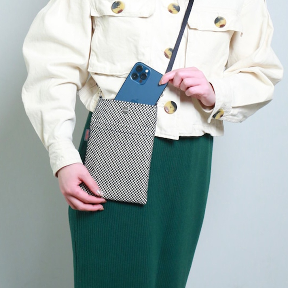一款黑色與白色棋盤格圖案的三河木綿刺子織手機包，由一位身穿米白色外套和綠色裙子的女士斜挎展示，手機包內裝有一部藍色手機。