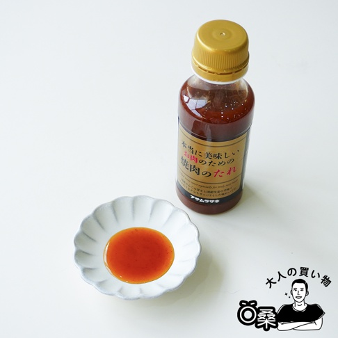 一瓶帶有金色瓶蓋的朝紫燒肉醬放置在白色桌面上，旁邊的小碟子盛裝著少量橘紅色的醬汁，右下角配有一個呈現雙臂交叉姿勢的人物插畫與相關文字標示。