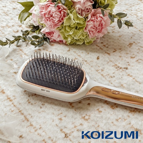 KOIZUMI正負離子養髮梳，白色機身搭配玫瑰金點綴，梳齒密集，上方擺放著粉色與綠色的花束。