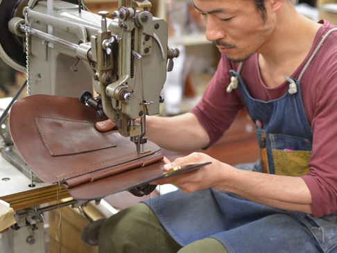 一位男性皮革匠人正在使用工業縫紉機縫製一個棕色皮革製成的迷你後背包。皮革表面有縫線細節，其中一塊皮革上有一個小口袋。