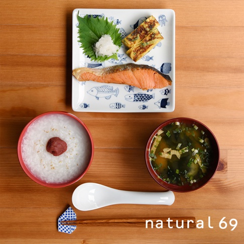 裝有烤鮭魚、玉子燒、蘿蔔泥和紫蘇葉的白色方形餐盤，盤面印有藍色魚圖案。旁邊是裝有飯的紅色碗和味噌湯的深紅色碗，以及白色湯匙和木筷。