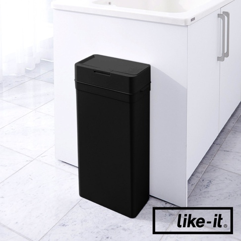 一個黑色、長方形的按壓式垃圾桶，帶有密封蓋，放置在白色櫥櫃旁的地板上。