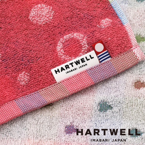 這張照片展示了兩條 Hartwell 今治產毛巾的特寫，上方為粉紅色底配白色圓點圖案的毛巾，邊緣有細緻的織帶，並縫有印著 Hartwell Imabari Japan 字樣的布標以及圓形標誌。下方露出部分為帶有彩色小象圖案的白色毛巾，畫面右下方同樣印有 Hartwell Imabari Japan 的黑色字樣，呈現出柔軟厚實的毛巾布質感。