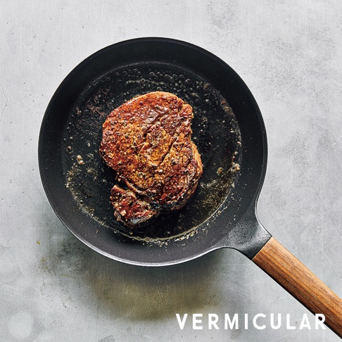 黑色琺瑯鑄鐵平底鍋，鍋內煎著一塊帶有香料的牛排，鍋柄為木質。