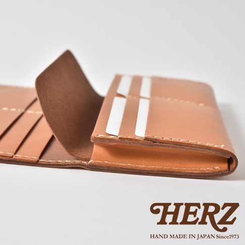 這是一張日本HERZ品牌職人手作皮革長夾的特寫照片。畫面採用側視角度，展示了淺棕色真皮皮夾開啟後的內部結構，可以看到精細的縫線工藝、層次分明的卡片夾層以及皮革厚實的邊緣細節，右下角印有HERZ品牌名稱與標語。
