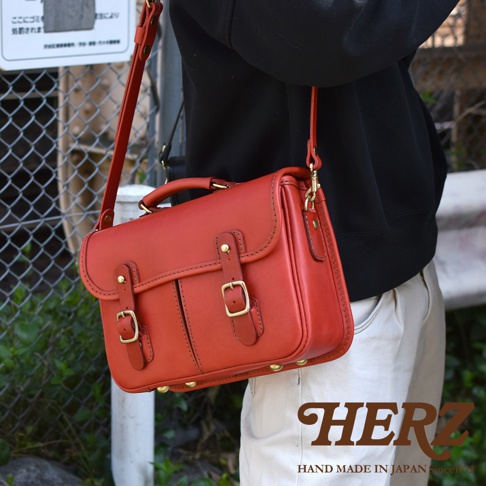 紅色皮革製成的雙用單肩包，有提把和可拆卸肩帶，包身有雙皮帶釦和金屬腳釘裝飾。