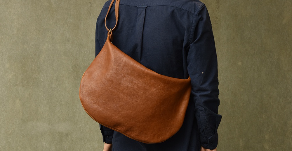 棕褐色皮革製成的半月形單肩包，斜挎於身著深藍色襯衫的人身上，包身線條柔和，材質紋理清晰可見。