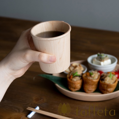 手持楓木寬口杯，杯身呈現天然淺木色，紋理清晰。杯中盛有深色液體。背景是木質桌面，擺放著日式料理，包括豆皮壽司、白玉豆腐和青菜。