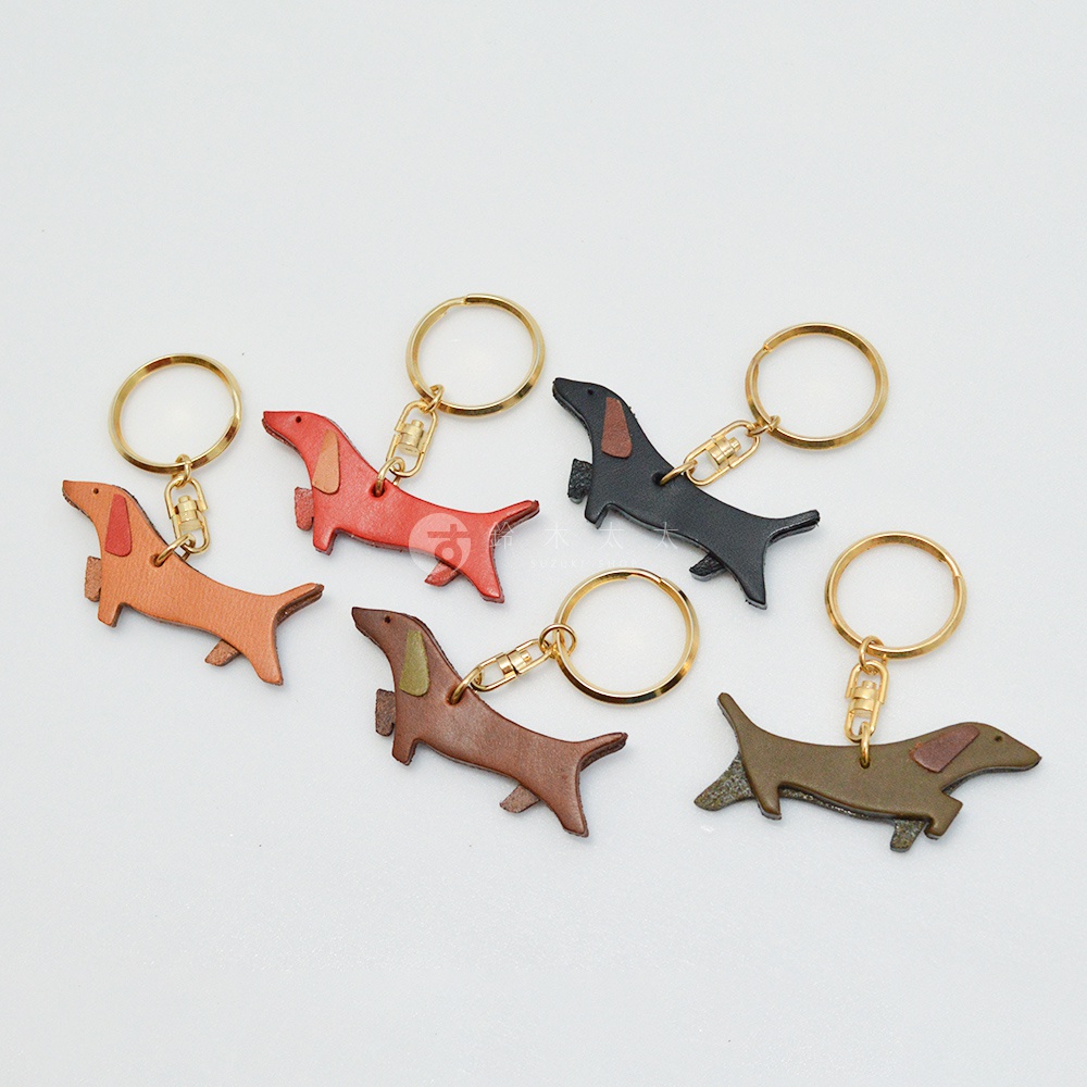 皮革狗狗造型鑰匙圈，共有橘棕、橘紅、黑、棕、綠棕五種顏色，以簡約線條呈現臘腸狗造型，搭配金色鑰匙圈。