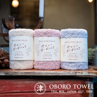 三條捲起的有機棉毛巾，依序為米白色、淺粉色與淺藍色，毛巾質地蓬鬆柔軟，包裝紙帶上有日文說明與棉花圖案。