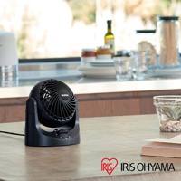 黑色桌上型空氣循環扇，扇葉網罩有螺旋紋路，底座有旋鈕開關，放置於木紋檯面上，背景為模糊的廚房窗戶與餐具。