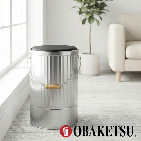 OBAKETSU 品牌手工製作的鍍鋅鋼儲物桶，頂部配有黑色坐墊，可兼作凳子，一旁是室內植物和沙發。
