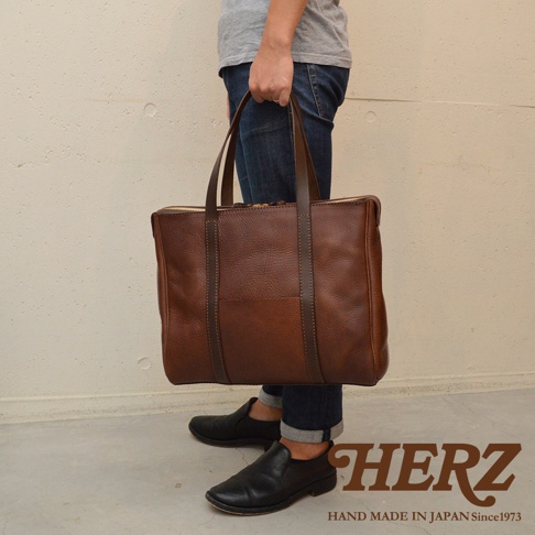 一位人士手提棕色皮革製的托特包，包款有拉鍊開合及雙提把，側面可見品牌標誌。