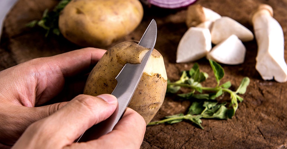 雙手正用削皮刀削切馬鈴薯，旁邊散落著切塊的菇類和綠色香草。