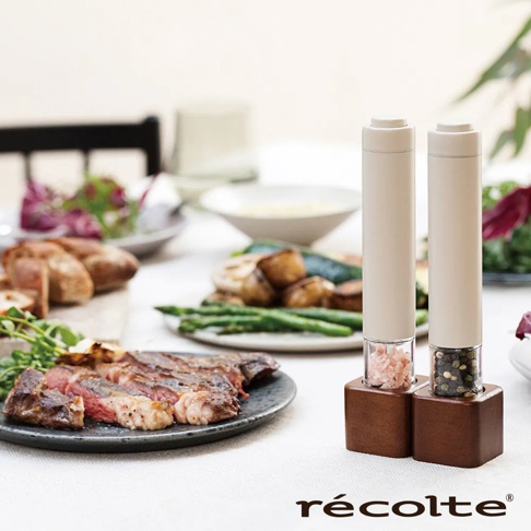兩支Récolte RMM-1電動研磨器，一支裝有粉紅色岩鹽，另一支裝有黑胡椒粒，均置於木製底座上，背景為烤牛排、烤蔬菜和沙拉。