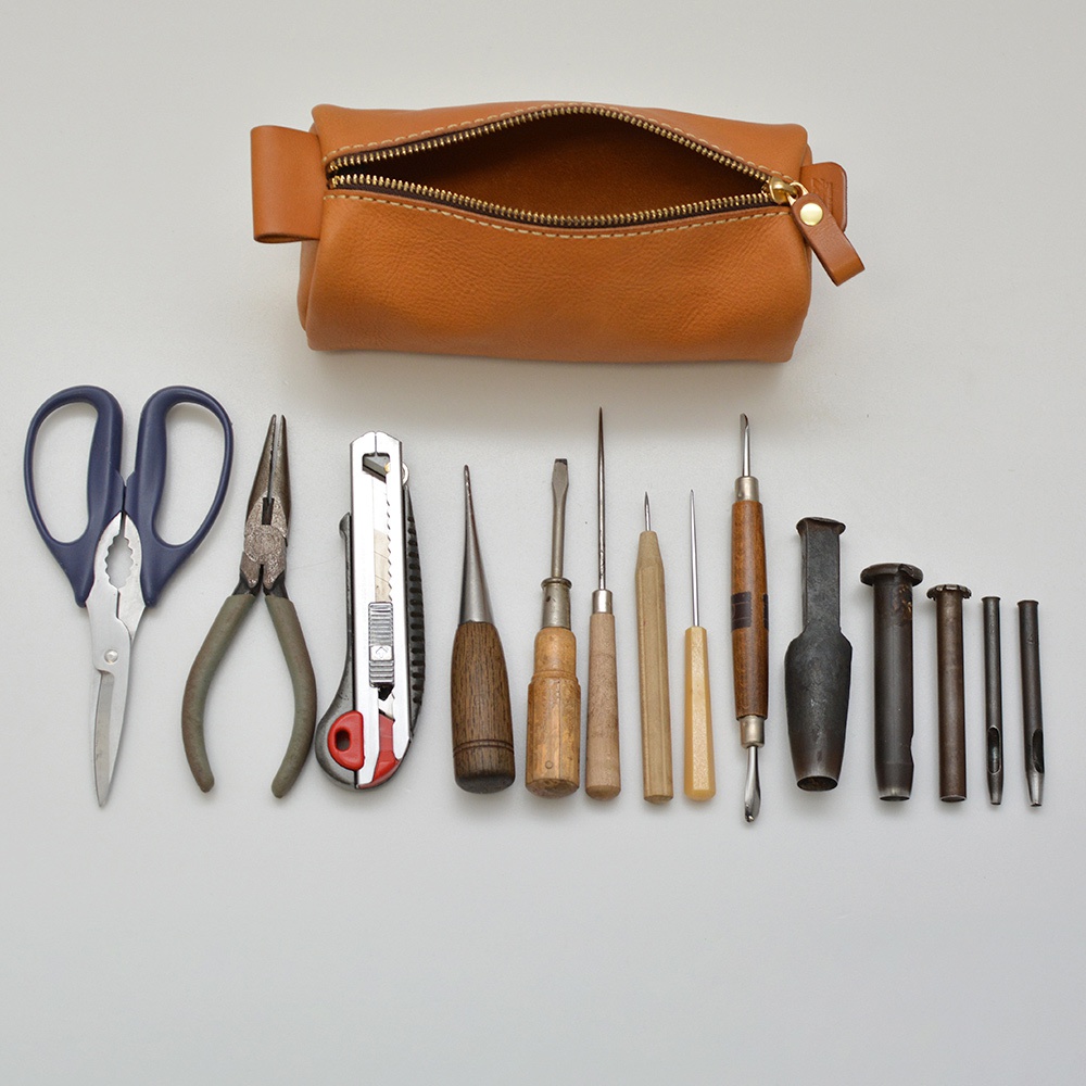 職人手作皮革多用途收納筆袋，淺棕色皮革材質，拉鍊開口，內含剪刀、鉗子、美工刀、各式皮革工具（如鑽孔器、壓痕筆、縫針等）。