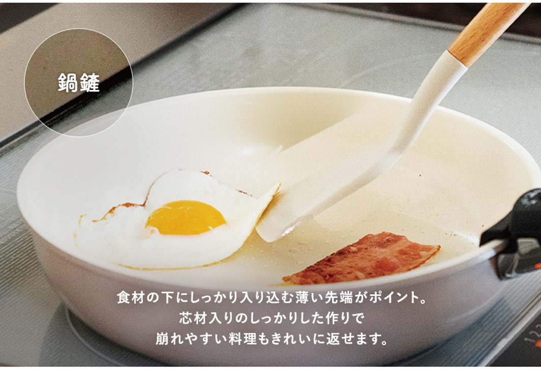 白色陶瓷鍋中煎著一顆太陽蛋和一片培根，一把白色鍋鏟（木質握柄）正要翻動食材。鍋邊緣有「鍋鏟」字樣的圓形標示。