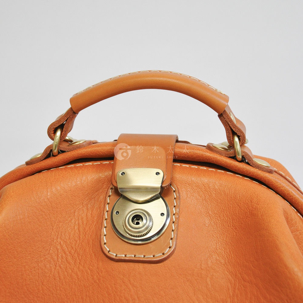 職人手作皮革輕旅行後背包 R-144，特寫展示其棕褐色皮革材質、金色旋轉鎖扣及縫線細節，以及提把的皮革與金屬連接處。