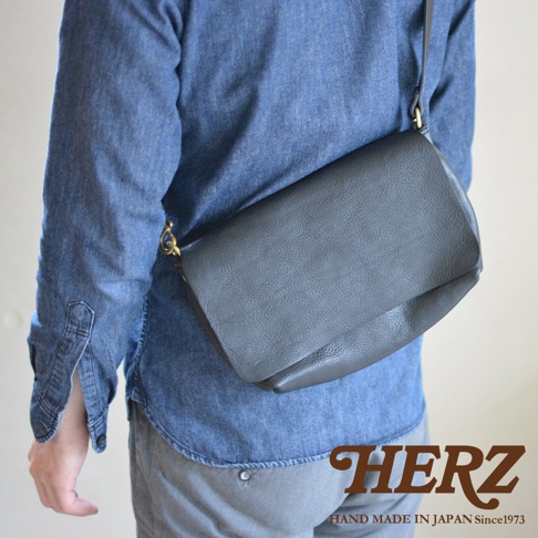 一個斜背著深灰色皮革包的男人，包包有著自然的紋理，搭配牛仔襯衫和灰色褲子。