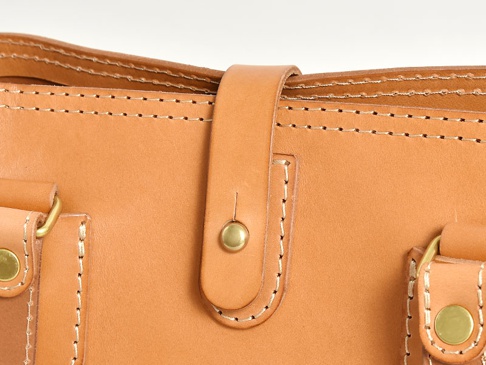 淺棕色皮革托特包局部特寫，展示精緻縫線、皮革翻蓋及黃銅色原子釦，材質為天然皮革。