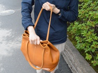 一位女士手持一個棕色皮革托特包，包身有縫線細節，搭配深藍色上衣和卡其色褲子，背景為綠色植物和石牆。