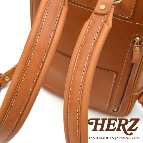 棕色皮革後背包的特寫，展示了兩條縫線精緻的皮革肩帶，並帶有金色金屬扣環。皮革表面光滑，質感細膩，整體呈現復古風格。