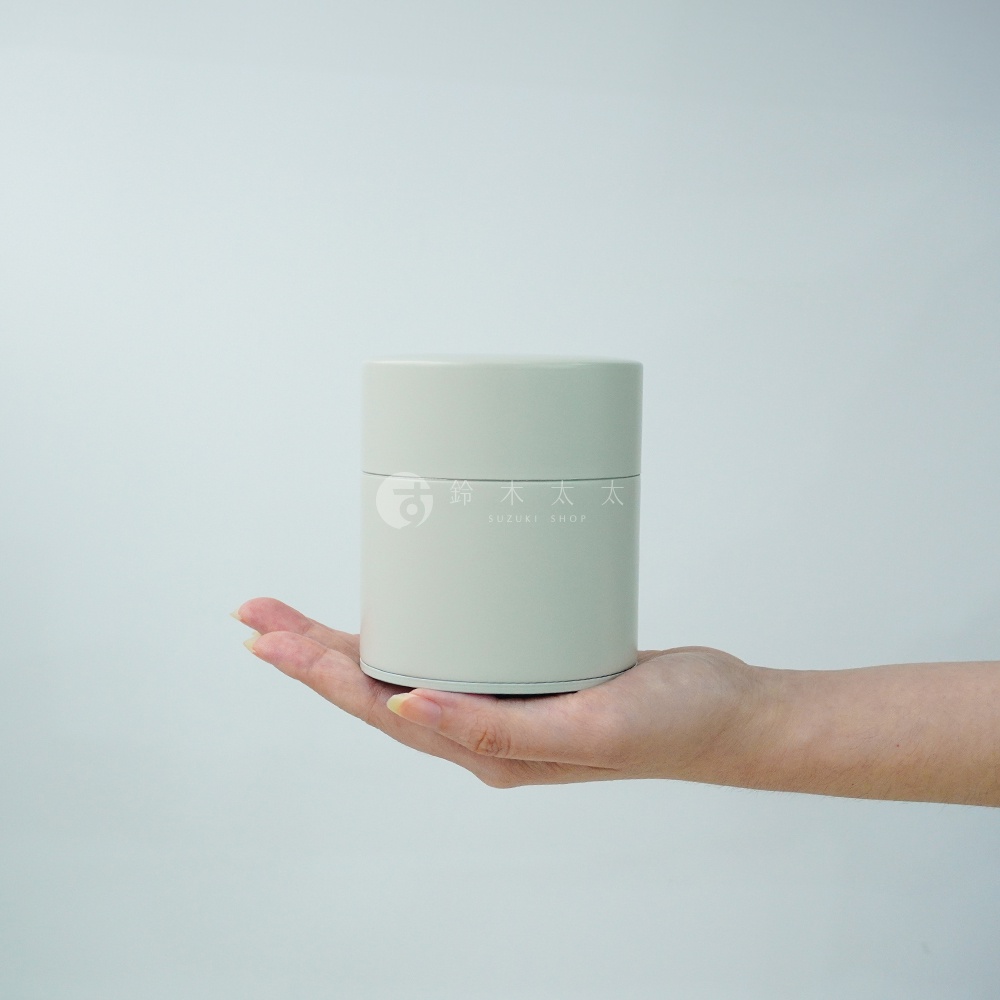 一只手托著一個簡約的淺灰色圓柱形錫製茶葉罐，罐身上印有「鈴木太太」及「SUZUKI SHOP」字樣。