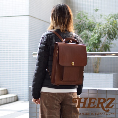 一位女性背著一個棕色皮革復古後背包，背包有頂部提把、翻蓋式主袋和前袋，配有金色鎖扣。