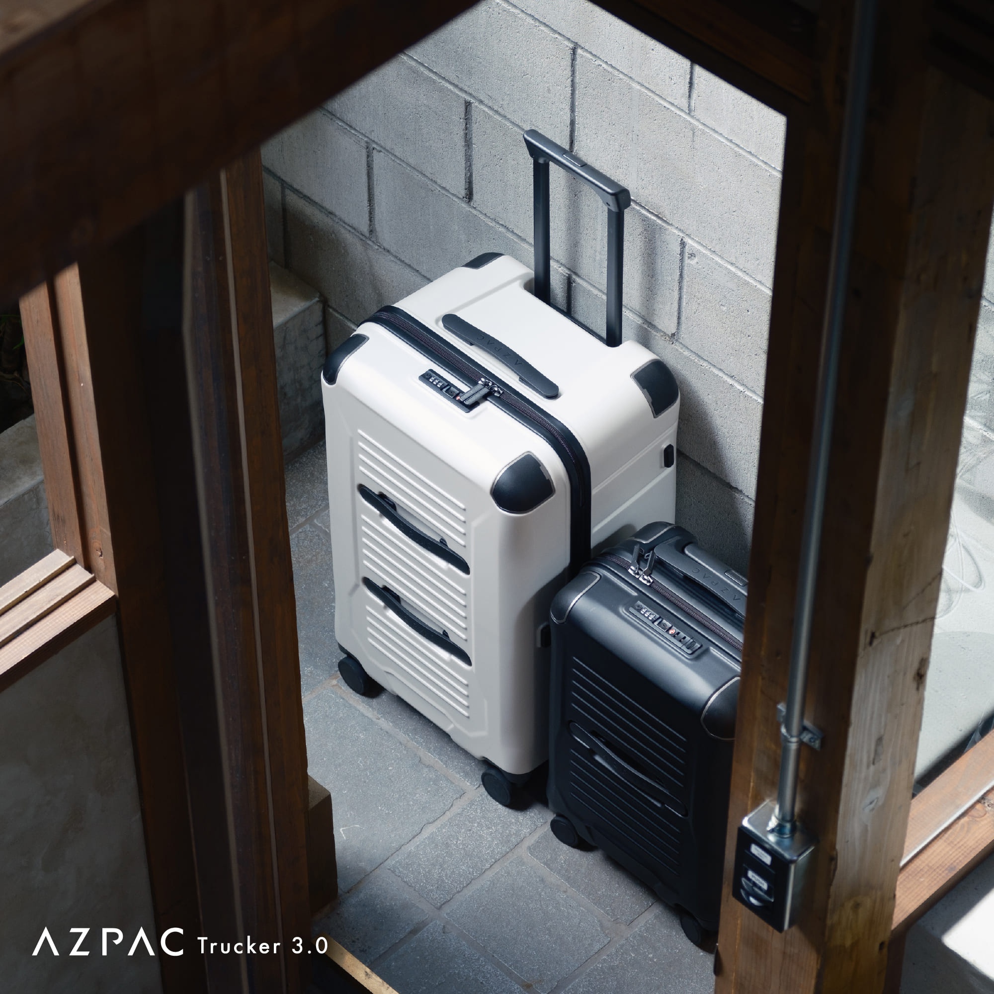 兩個一黑一白的 AZPAC Trucker 3.0 行李箱放置在室內水泥牆角邊，左側白色行李箱拉桿升起，右側黑色行李箱則較為小巧，兩者皆為硬殼設計且表面帶有橫向線條紋理，畫面呈現簡約的工業風格。