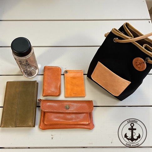 一組神戶clueto品牌皮件與束口包，包含一個棕色筆記本、一個橘色卡夾、一個橘色零錢包、一個橘色長夾，以及一個黑色尼龍搭配皮革裝飾的束口包。