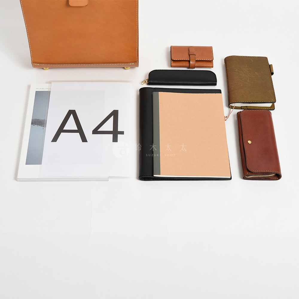 側邊擺放著多款皮革配件，包含一個棕色皮革卡夾，一個黑色拉鍊筆袋，一本裸棕色封面的筆記本，以及一本深綠色封面的小筆記本，旁邊還有一個棕色長夾。
